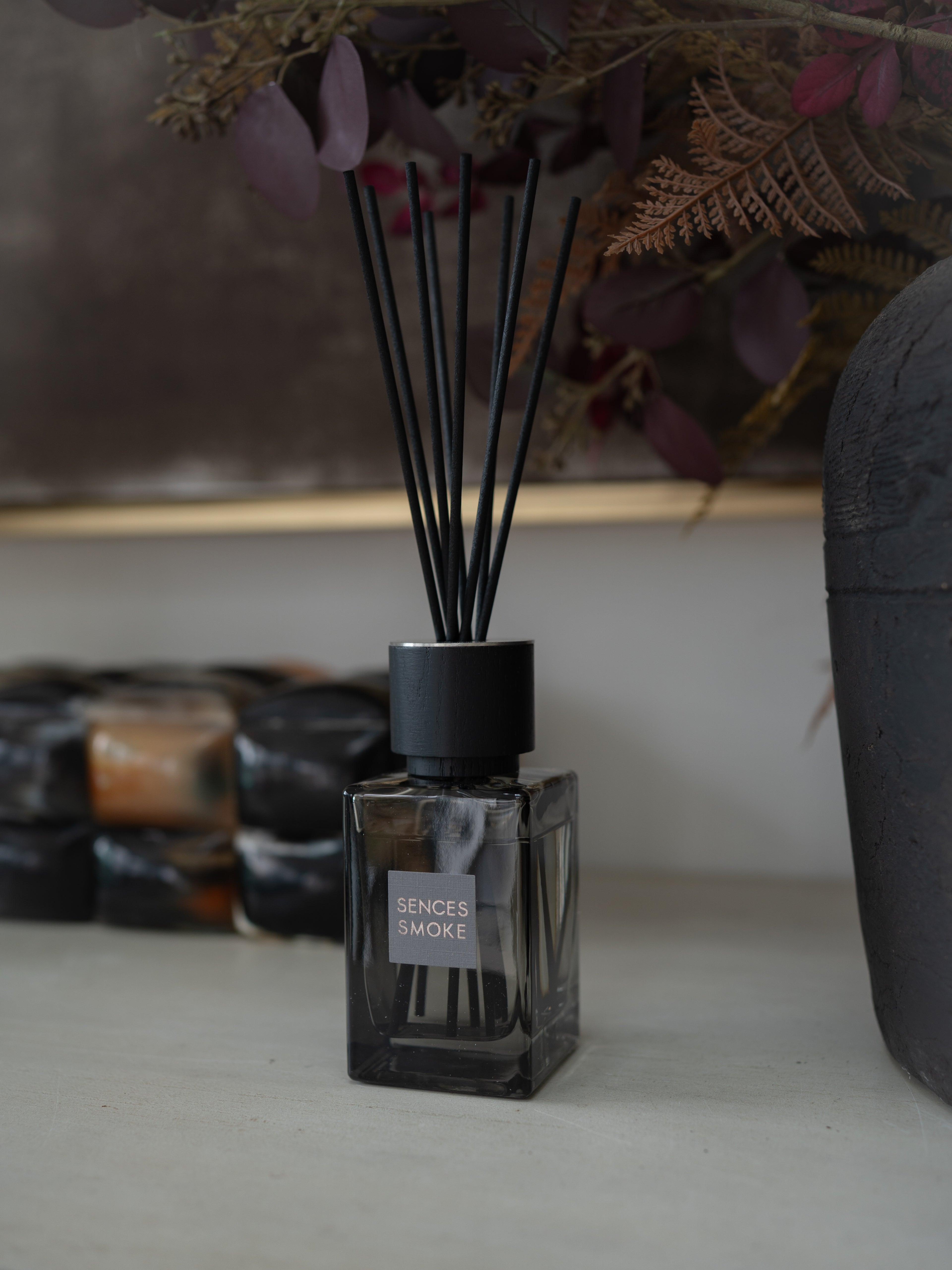 A sences Smoke reed diffuser in a dark square glass bottle with black reeds, styled on a tabletop with dried botanicals and other diffusers in the background.