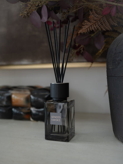 A sences Smoke reed diffuser in a dark square glass bottle with black reeds, styled on a tabletop with dried botanicals and other diffusers in the background.