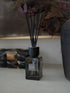 A sences Smoke reed diffuser in a dark square glass bottle with black reeds, styled on a tabletop with dried botanicals and other diffusers in the background.