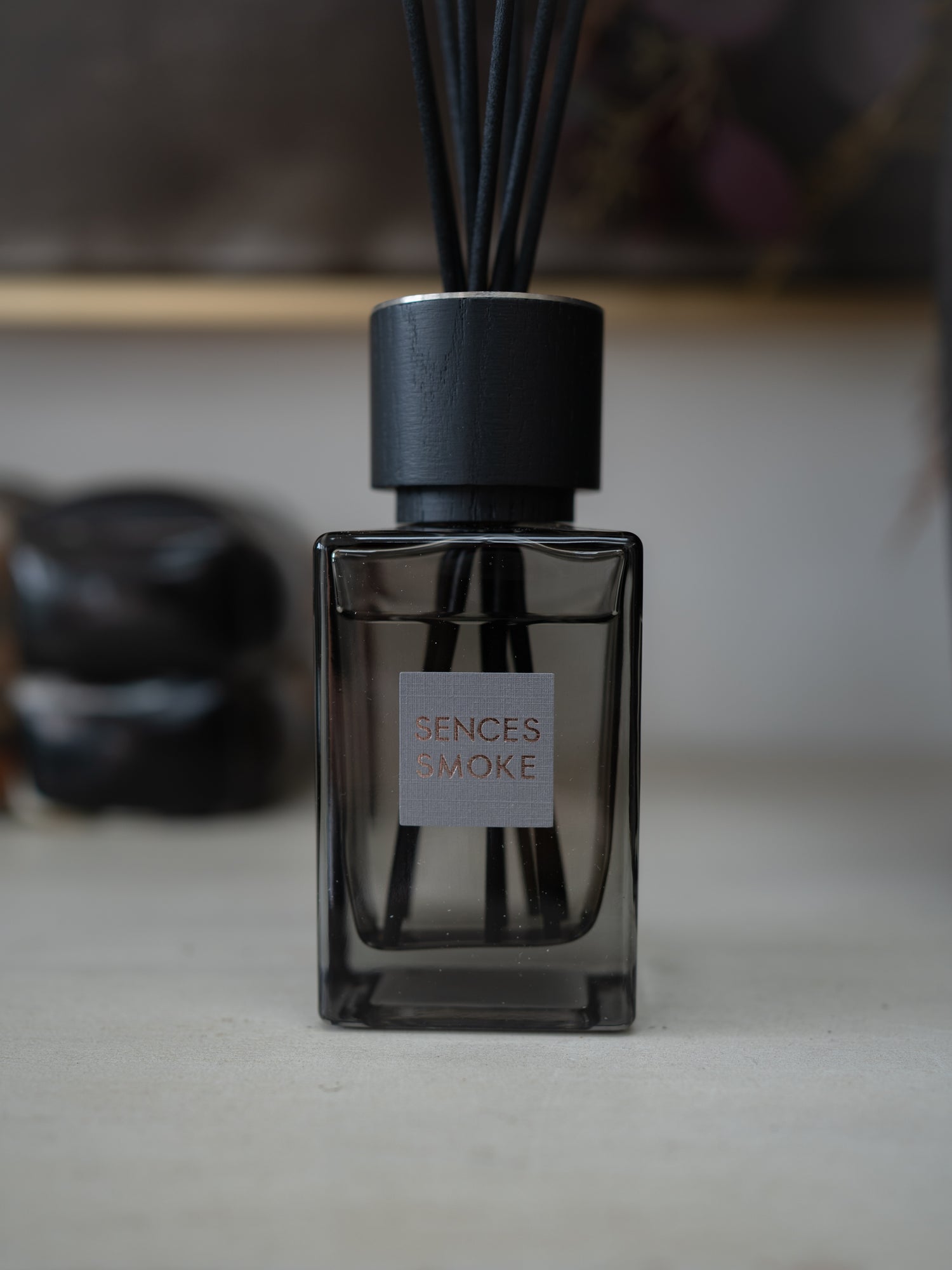 A reed diffuser bottle with a dark smoky glass body, black cap, and black reeds, featuring a square label that reads &