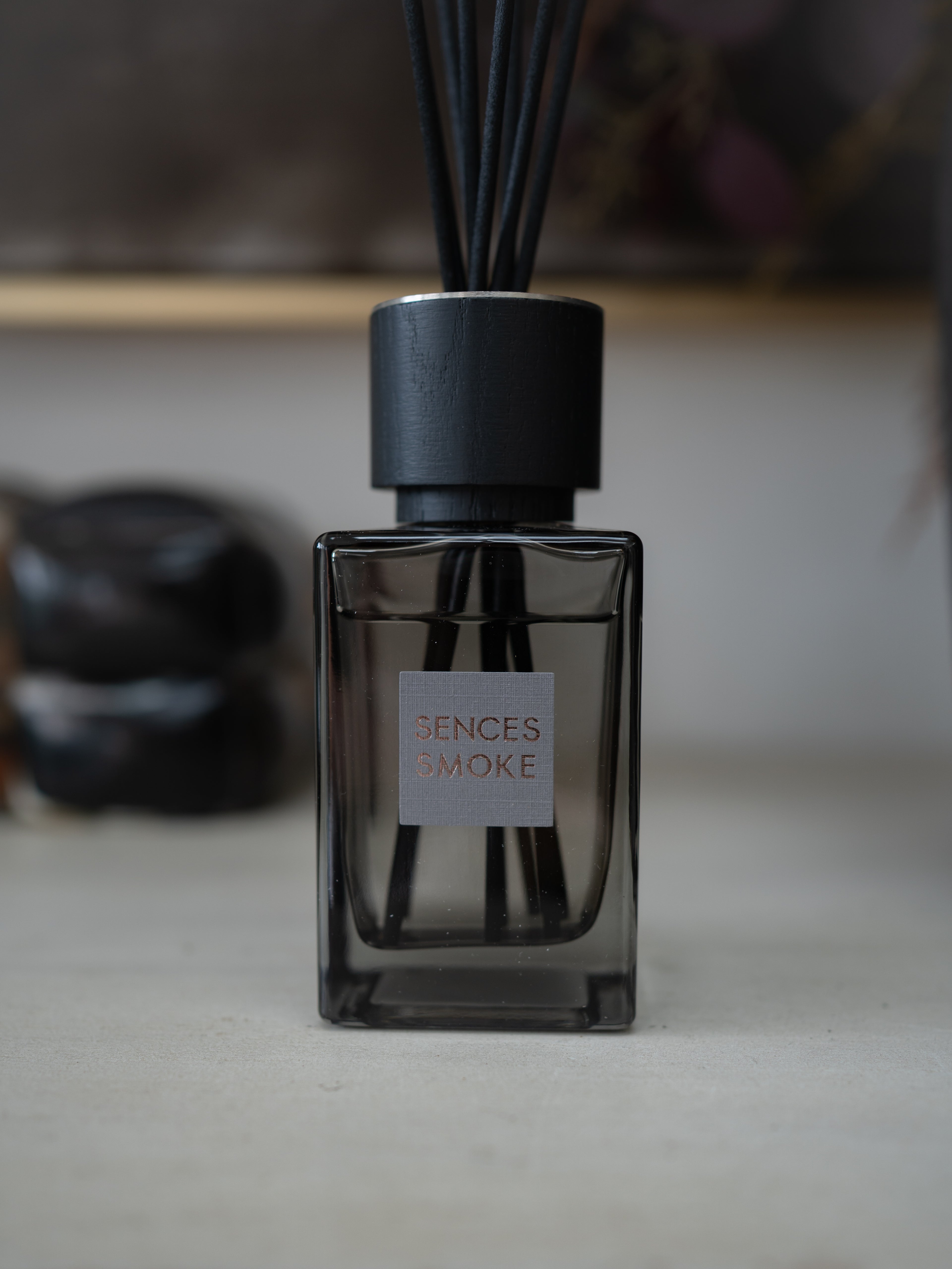 A reed diffuser bottle with a dark smoky glass body, black cap, and black reeds, featuring a square label that reads &