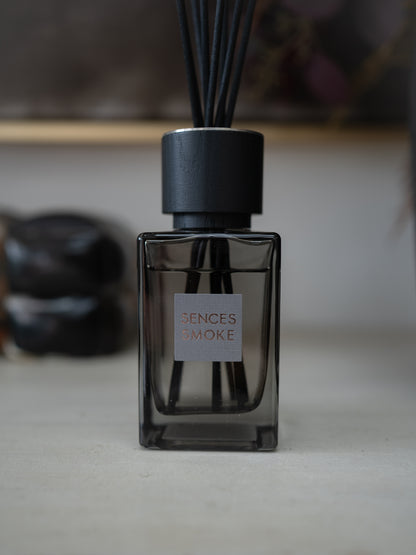 A reed diffuser bottle with a dark smoky glass body, black cap, and black reeds, featuring a square label that reads &
