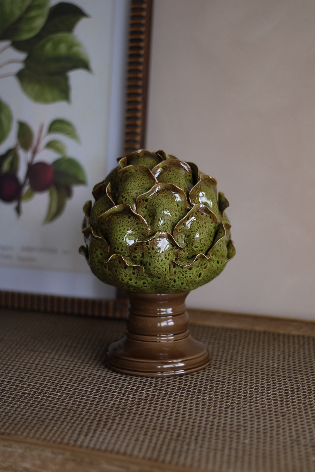 Decorative ceramic artichoke on a textured surface with a framed picture in the background.