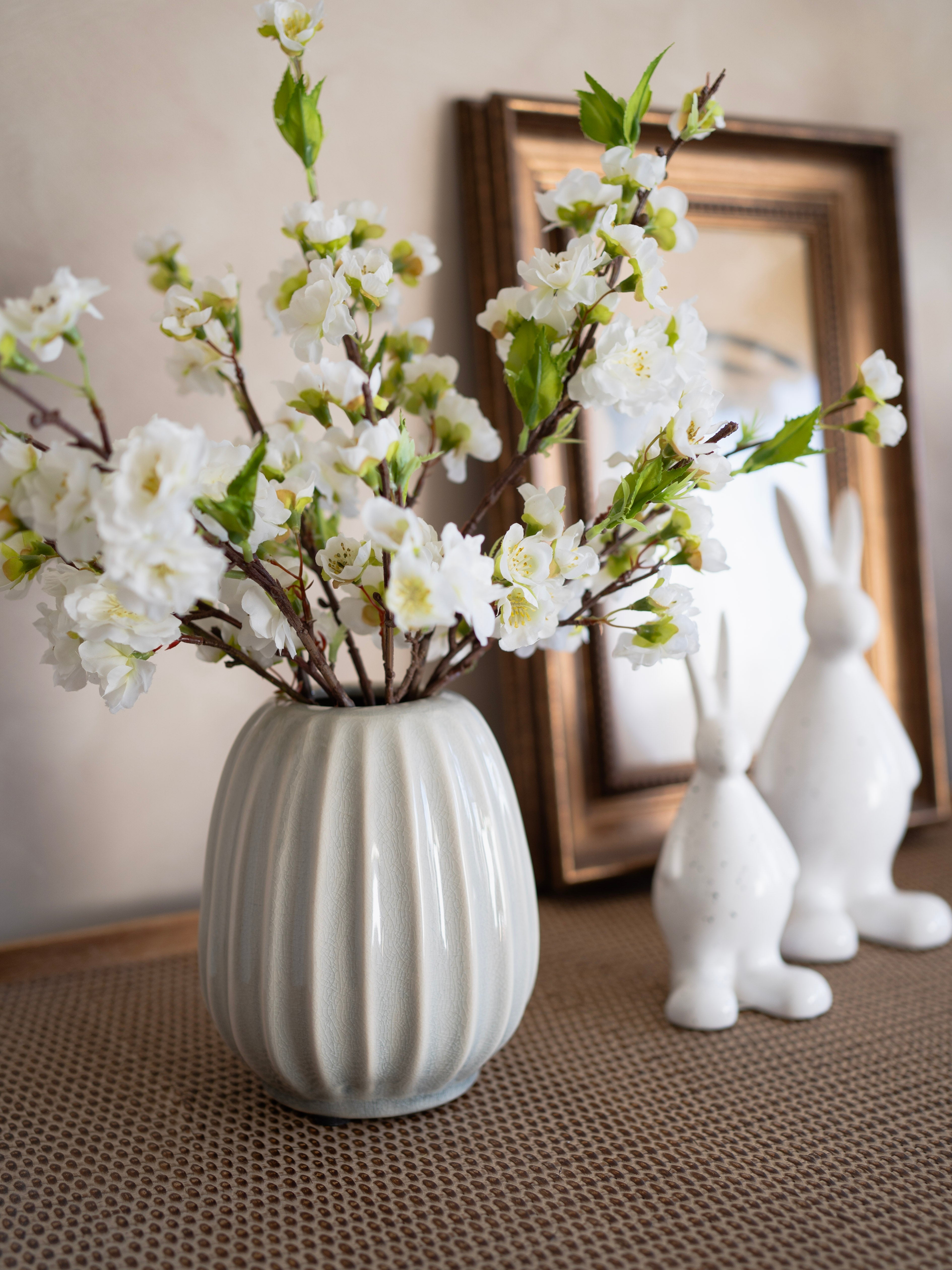 White vase with white flowers on a textured surface with decorative rabbits and a mirror in the background.
