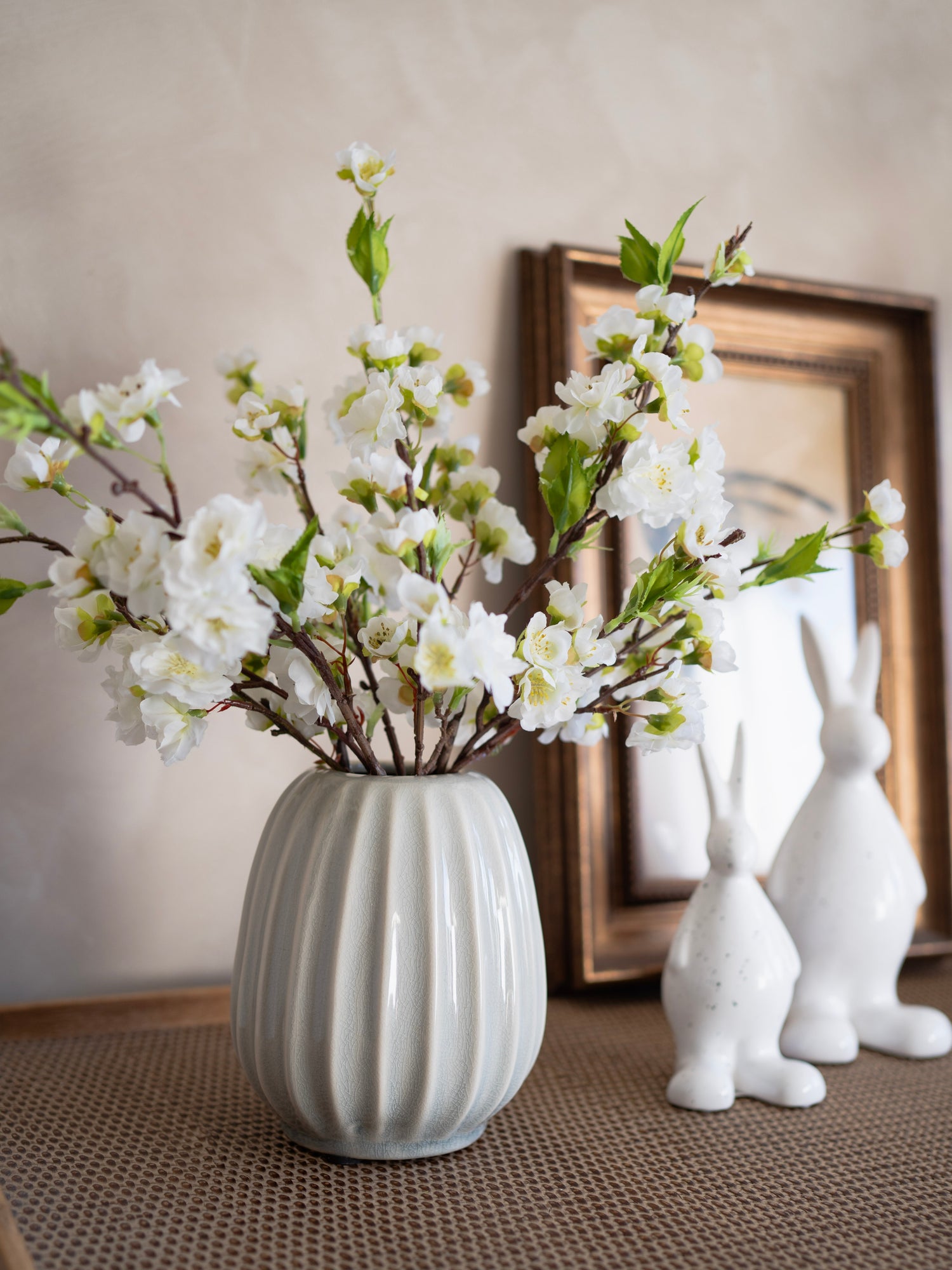 Grey ribbed vase with white flowers on a textured surface with decorative rabbits and a framed picture in the background.