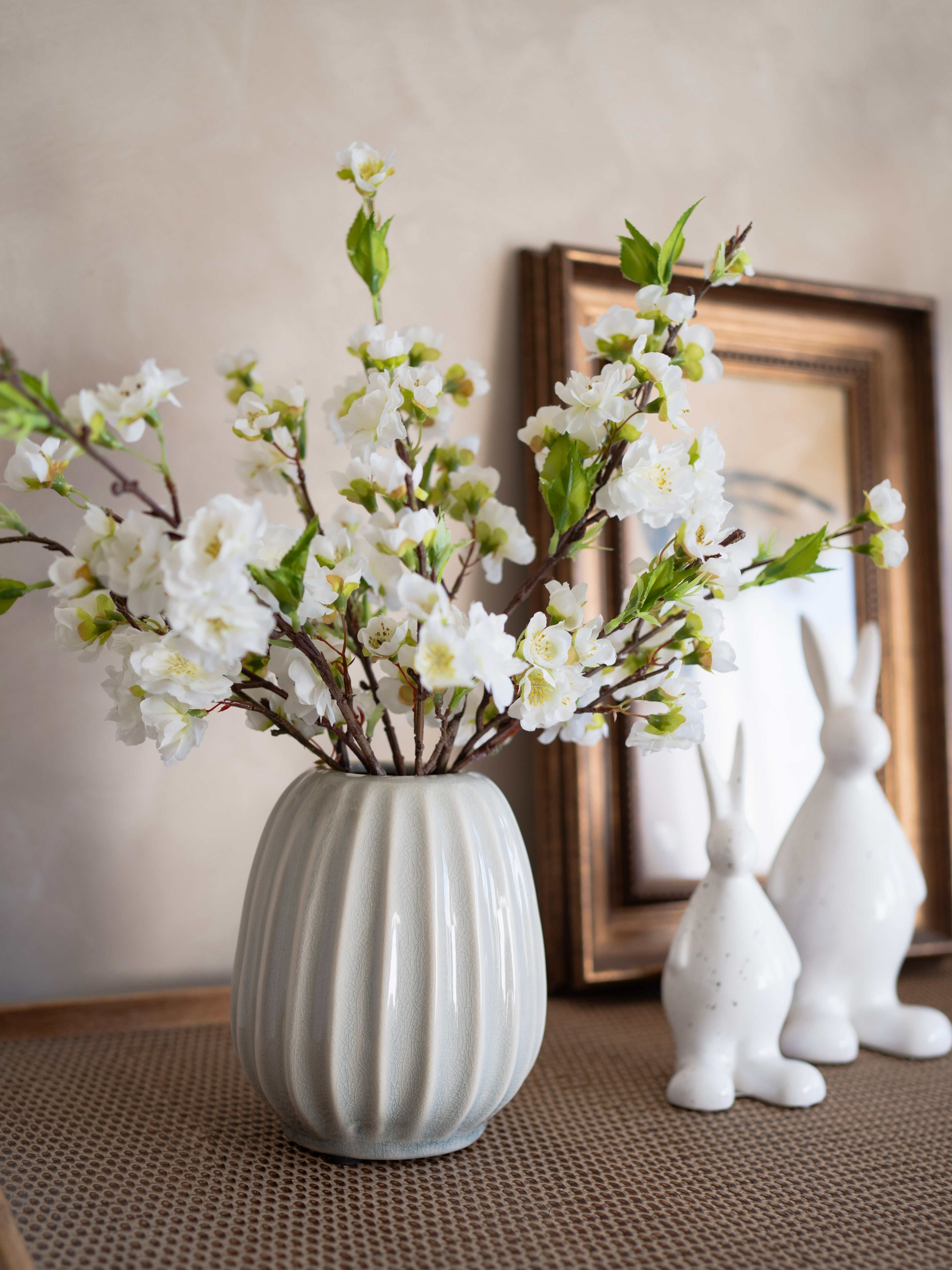 Grey ribbed vase with white flowers on a textured surface with decorative rabbits and a framed picture in the background.