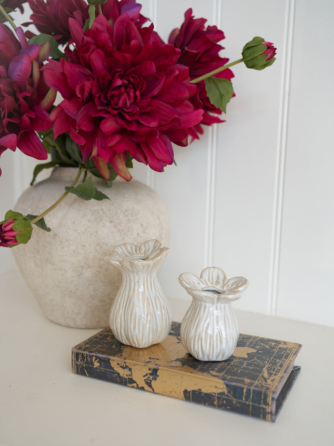 Decorative setting with a large vase of red flowers, two smaller vases, and a book on a white surface.