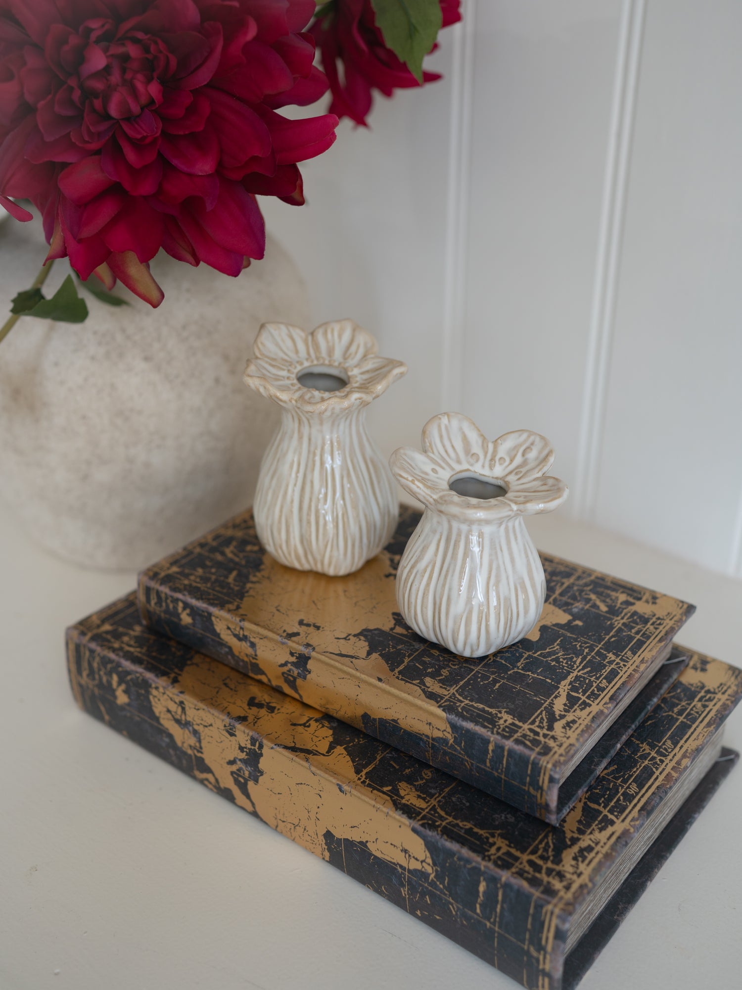 Decorative ceramic bud vases on vintage books with a vase of red flowers in the background.