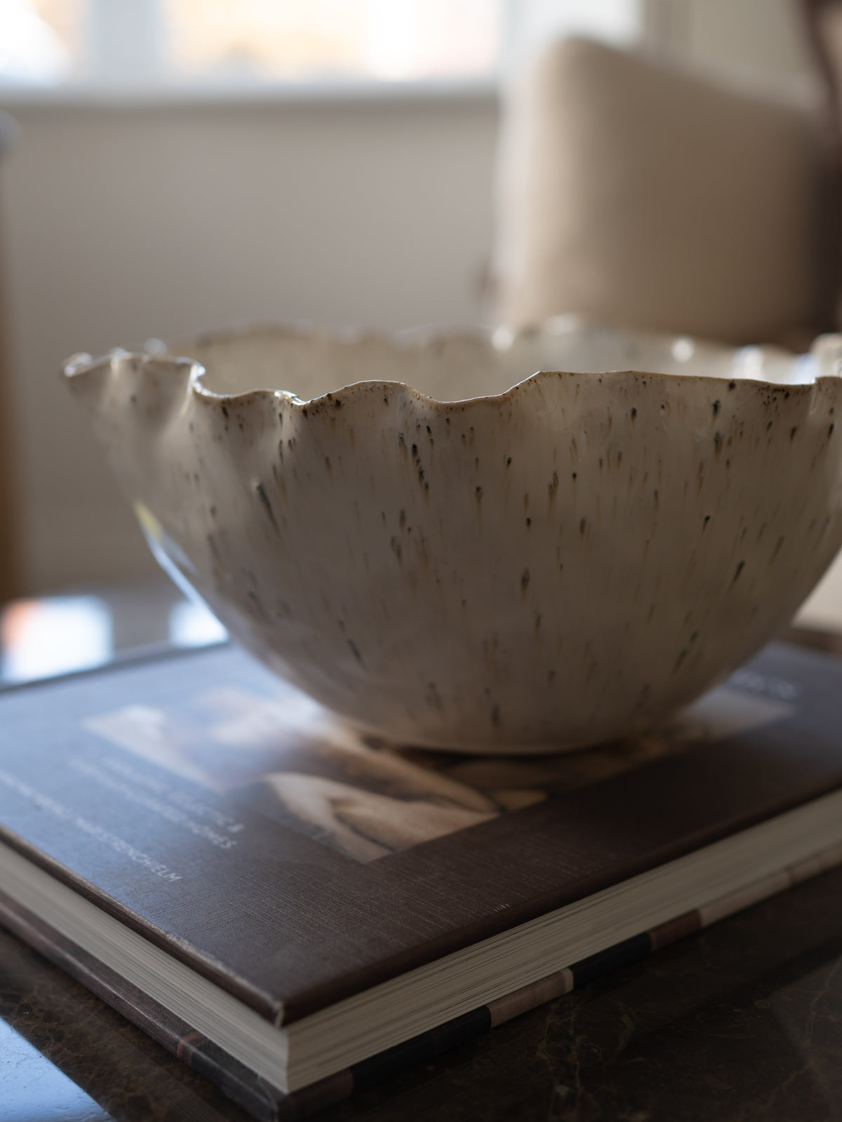A large, scalloped-edge ceramic bowl with a speckled cream and brown glaze, resting on a dark book in a softly lit indoor setting.