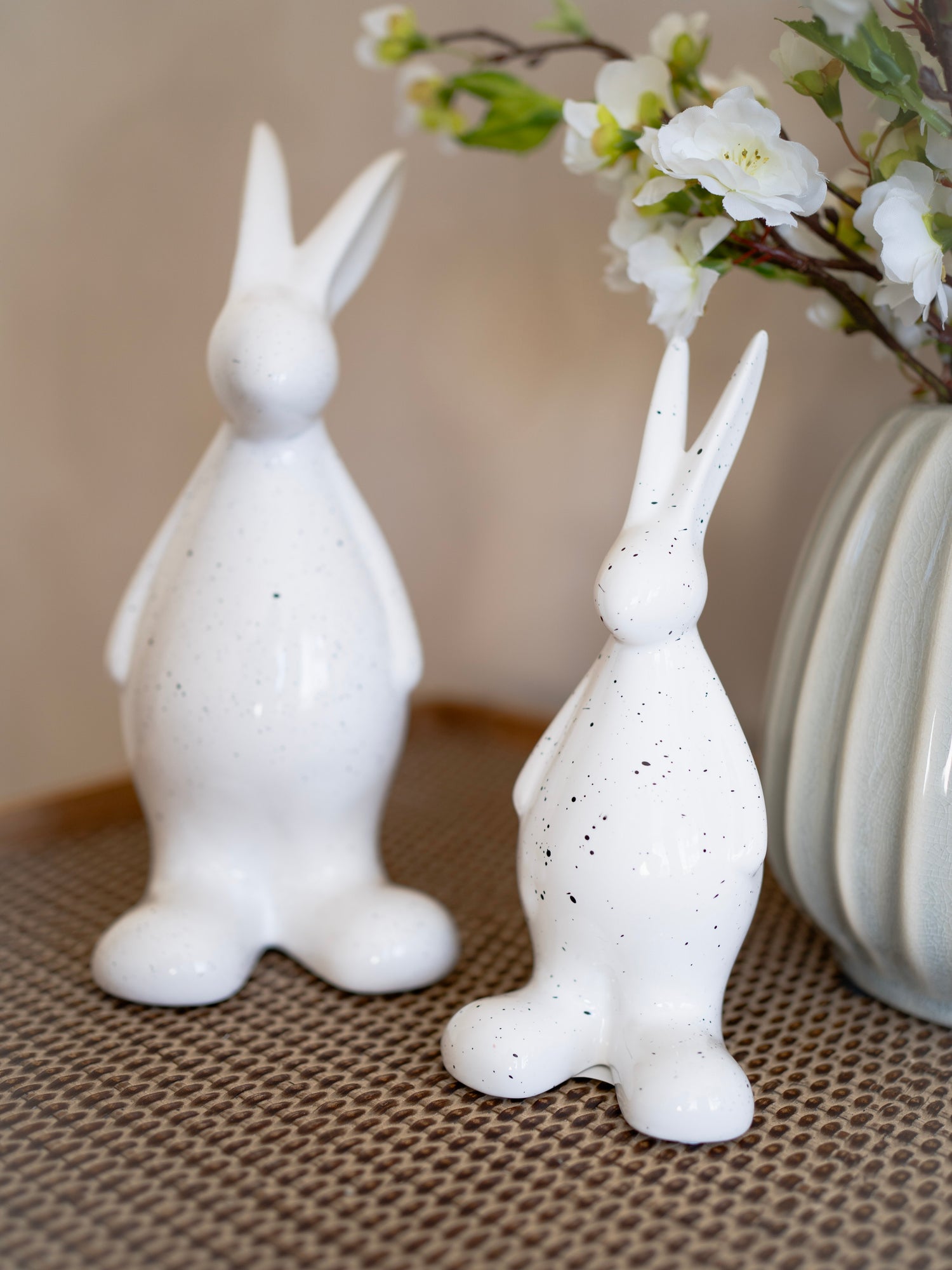 Two white ceramic rabbit figurines on a textured surface with a vase of flowers in the background.