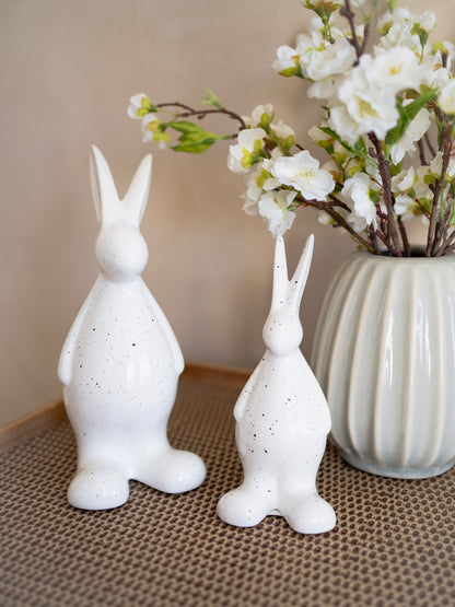 Two white ceramic rabbit sculptures on a textured surface with a vase of flowers in the background.