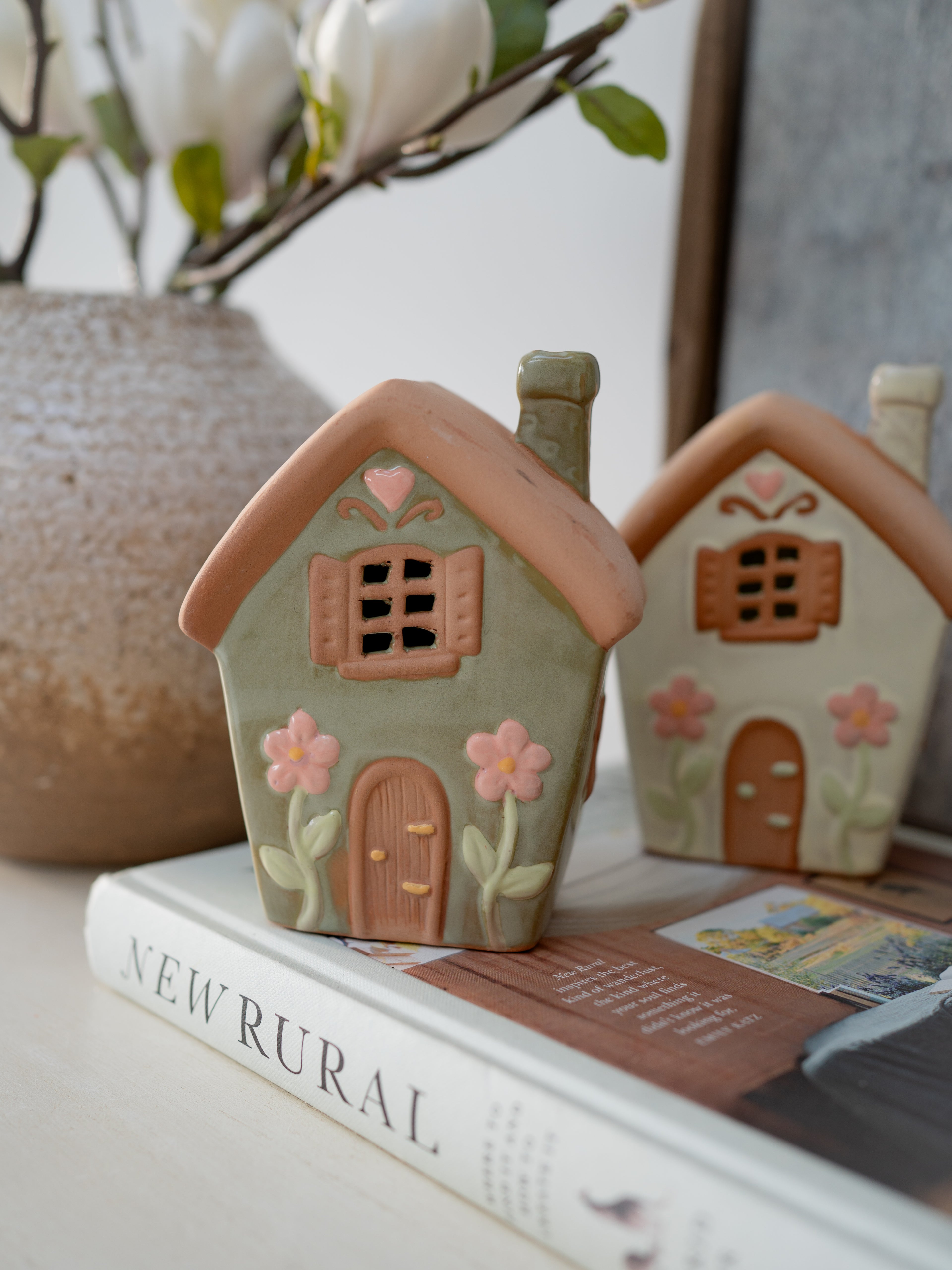 Two small ceramic houses with floral designs on a surface with books and a plant.