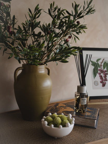 Decorative setting with a vase of greenery, a bowl of green balls, and a diffuser on a table.