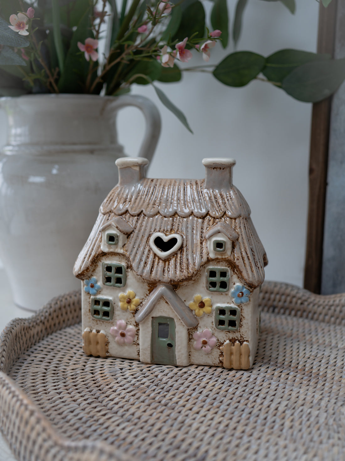 Small ceramic house with a heart window on a woven surface with a blurred vase and flowers in the background.