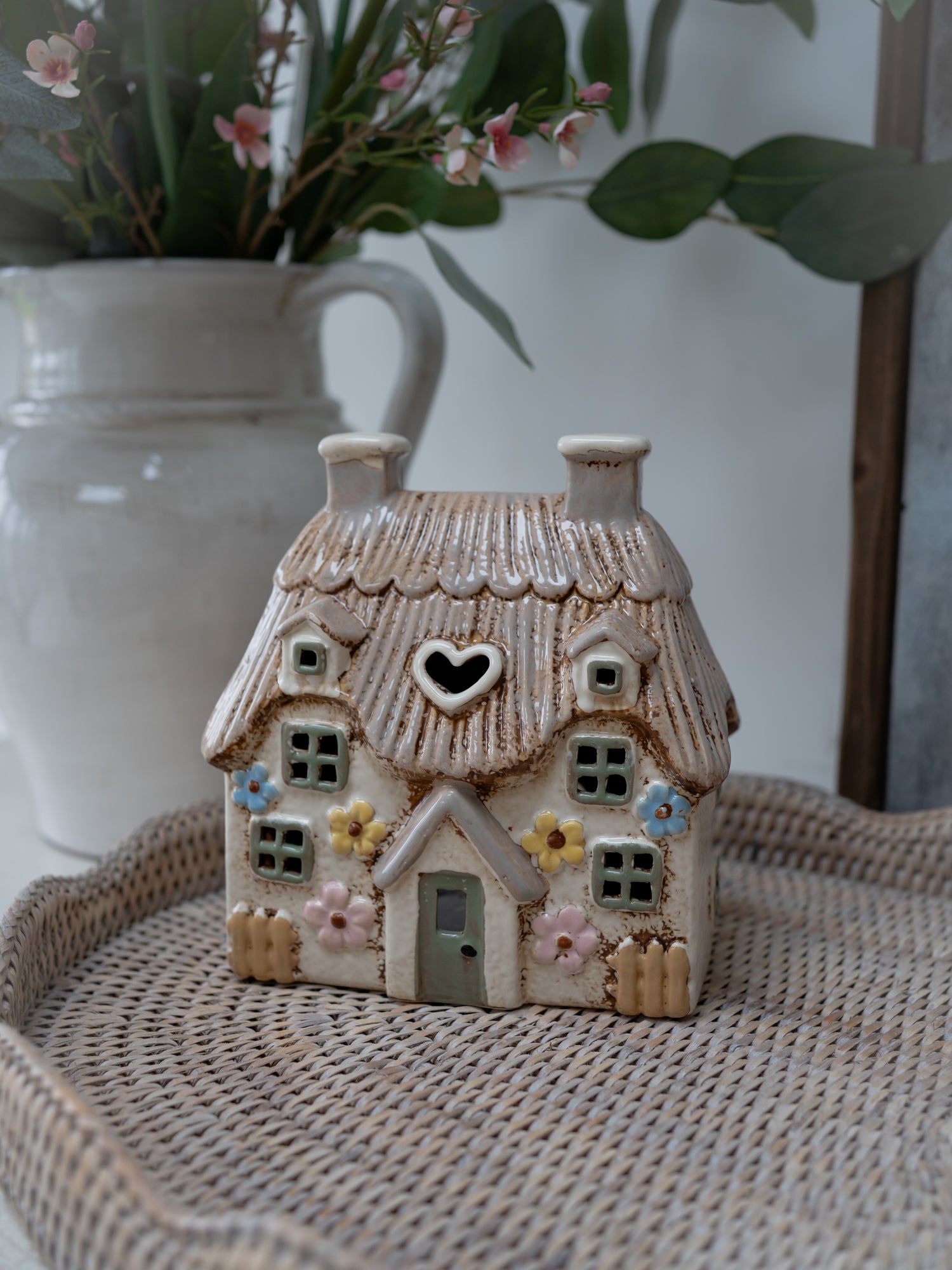 Small ceramic house with a heart window on a woven surface with a blurred vase and flowers in the background.
