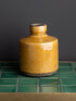 MUSTARD ceramic bottle on a green tiled surface with a dark gray background