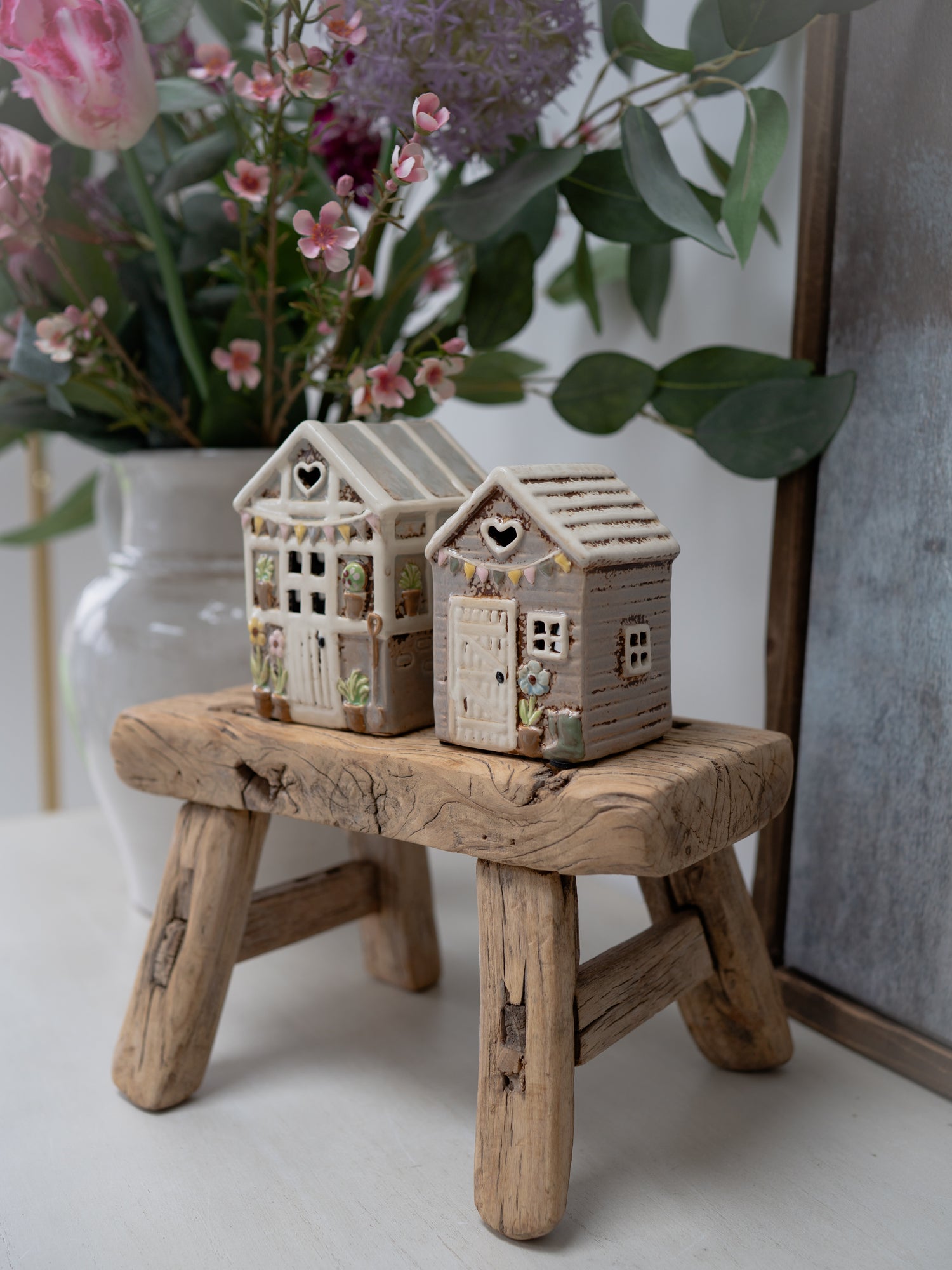 Two small ceramic houses on a wooden stool with flowers in the background