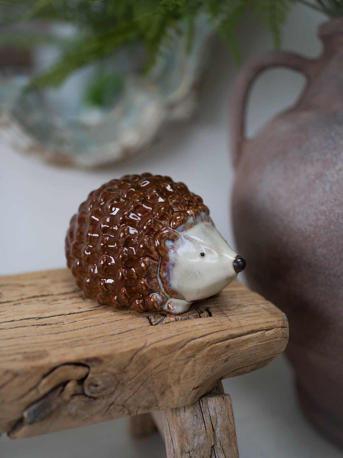 A small ceramic hedgehog figurine with a glossy brown textured body and cream-colored face, resting on a rustic wooden block with a blurred ceramic vase in the background.