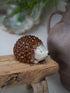 A small ceramic hedgehog figurine with a glossy brown textured body and cream-colored face, resting on a rustic wooden block with a blurred ceramic vase in the background.