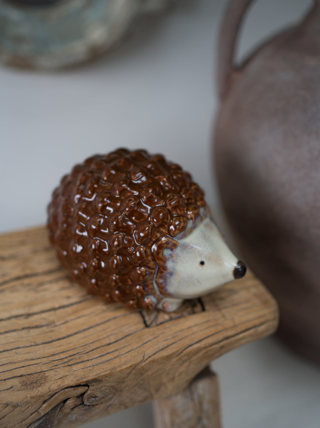A small ceramic hedgehog figurine with a glossy brown textured body and cream-colored face, resting on a rustic wooden block.
