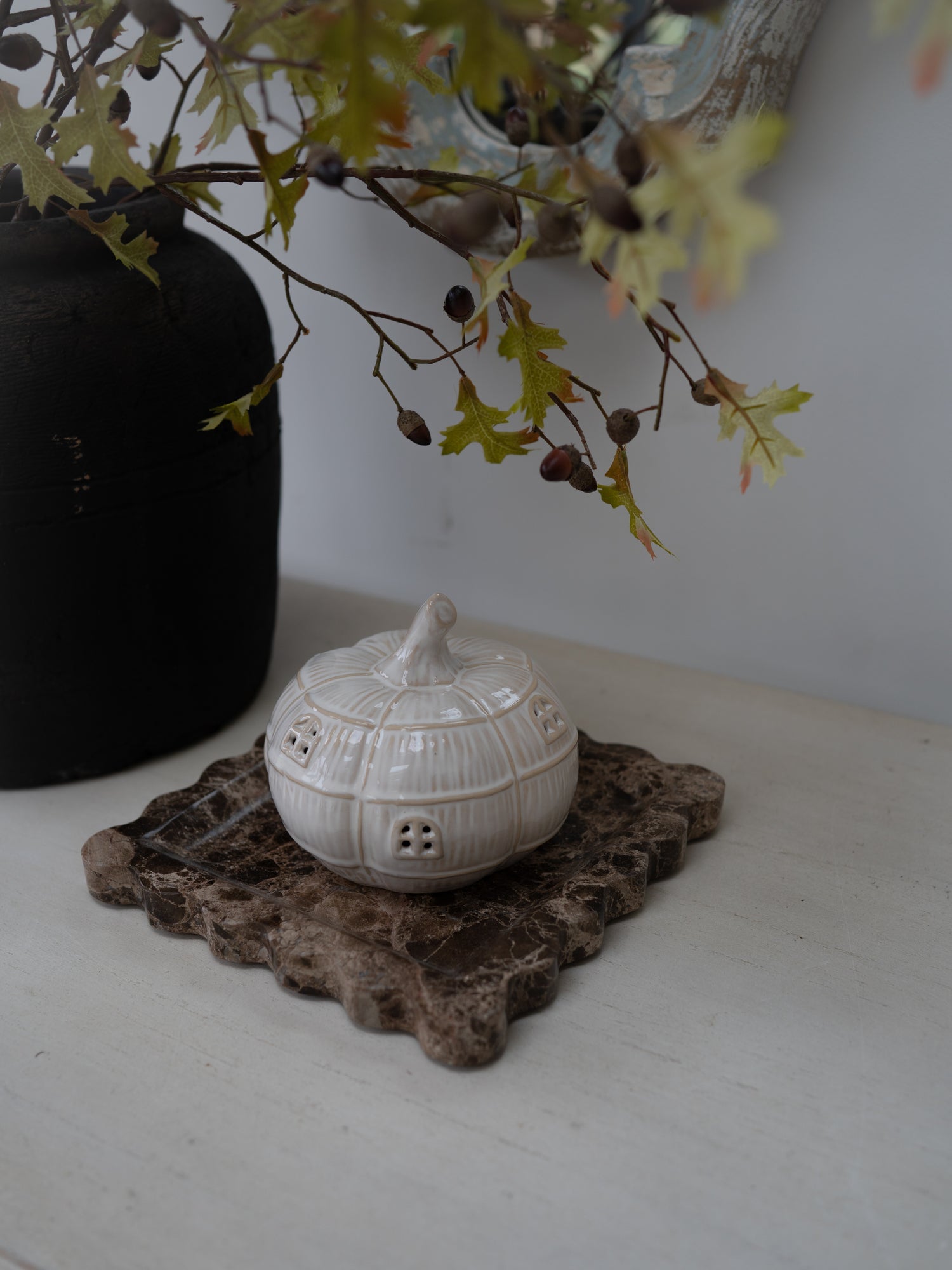 A small white ceramic house-shaped pumpkin sits on a rustic brown stone coaster on a white tabletop, with a black vase and leafy branches in the background.