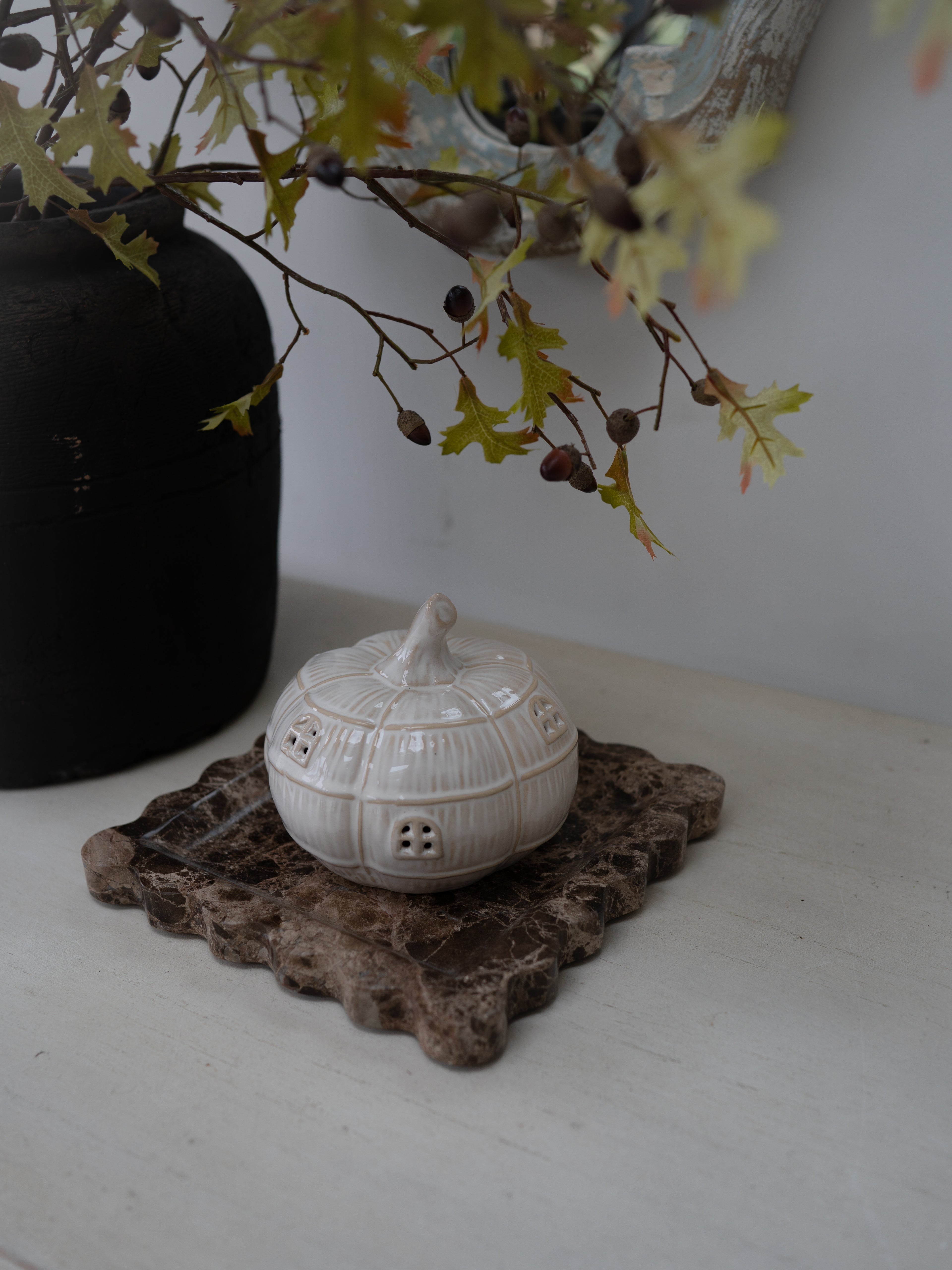 A small white ceramic house-shaped pumpkin sits on a rustic brown stone coaster on a white tabletop, with a black vase and leafy branches in the background.