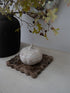A small white ceramic house-shaped pumpkin sits on a rustic brown stone coaster on a white tabletop, with a black vase and leafy branches in the background.
