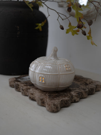 A small white ceramic pumpkin-shaped container with a lid and warm glowing windows, displayed on a rustic stone slab with a dark vase and hanging branches in the background.