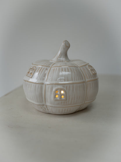 A small ceramic pumpkin-shaped house with a lit warm yellow window, shown on a neutral light gray background.