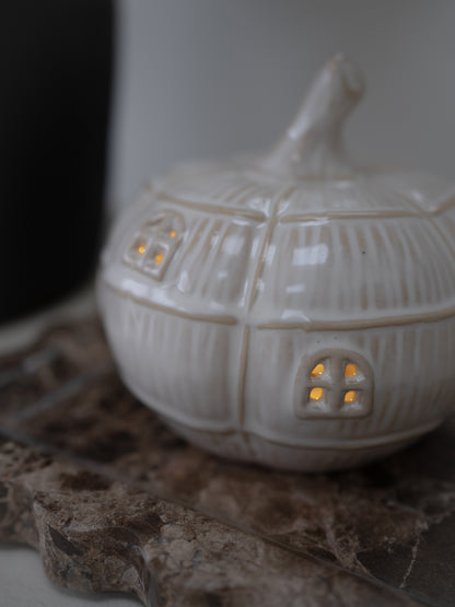 A small white ceramic house-shaped pumpkin led with ribbed texture and glowing warm yellow lights in the windows, resting on a textured stone surface.