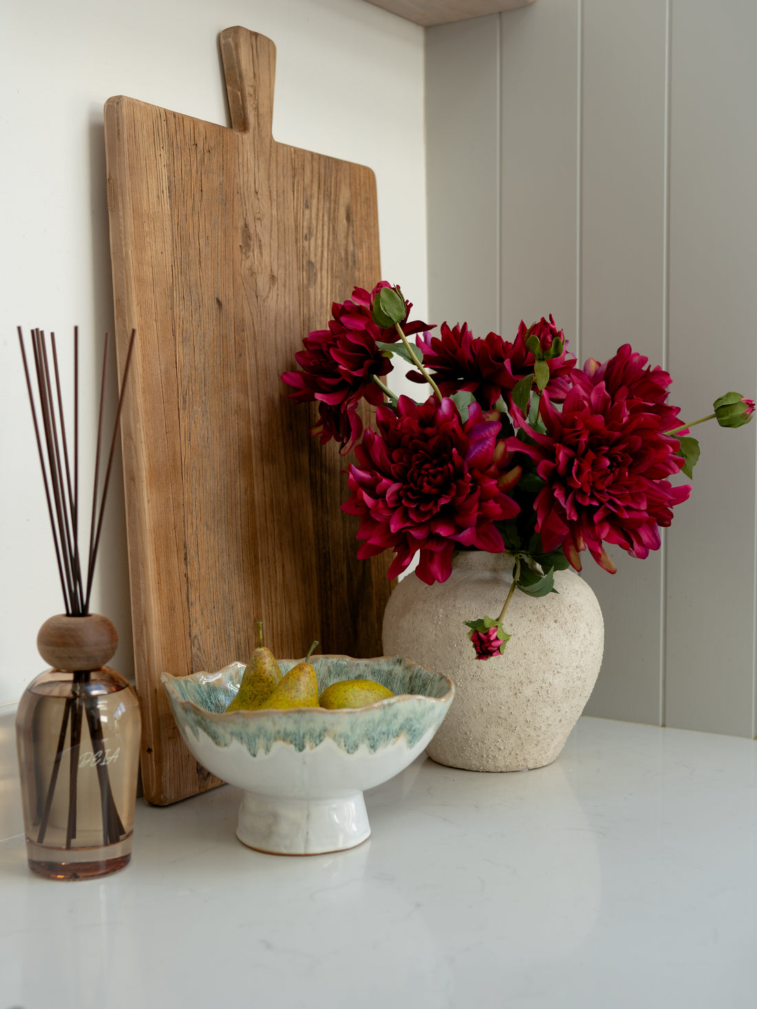 Decorative setting with a wooden cutting board, vase of red flowers, bowl with pears, and reed diffuser on a white surface.