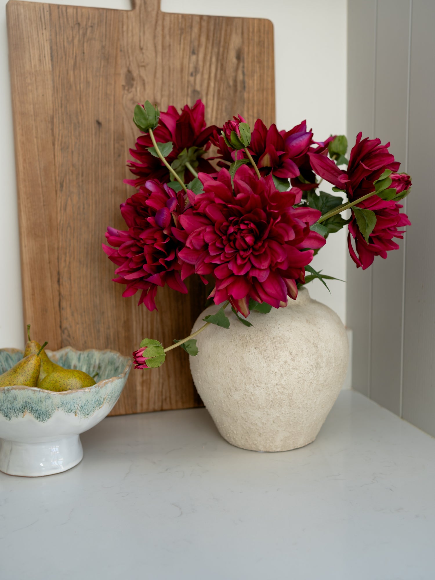 Vase with red flowers on a white surface next to a wooden cutting board and ceramic bowl.