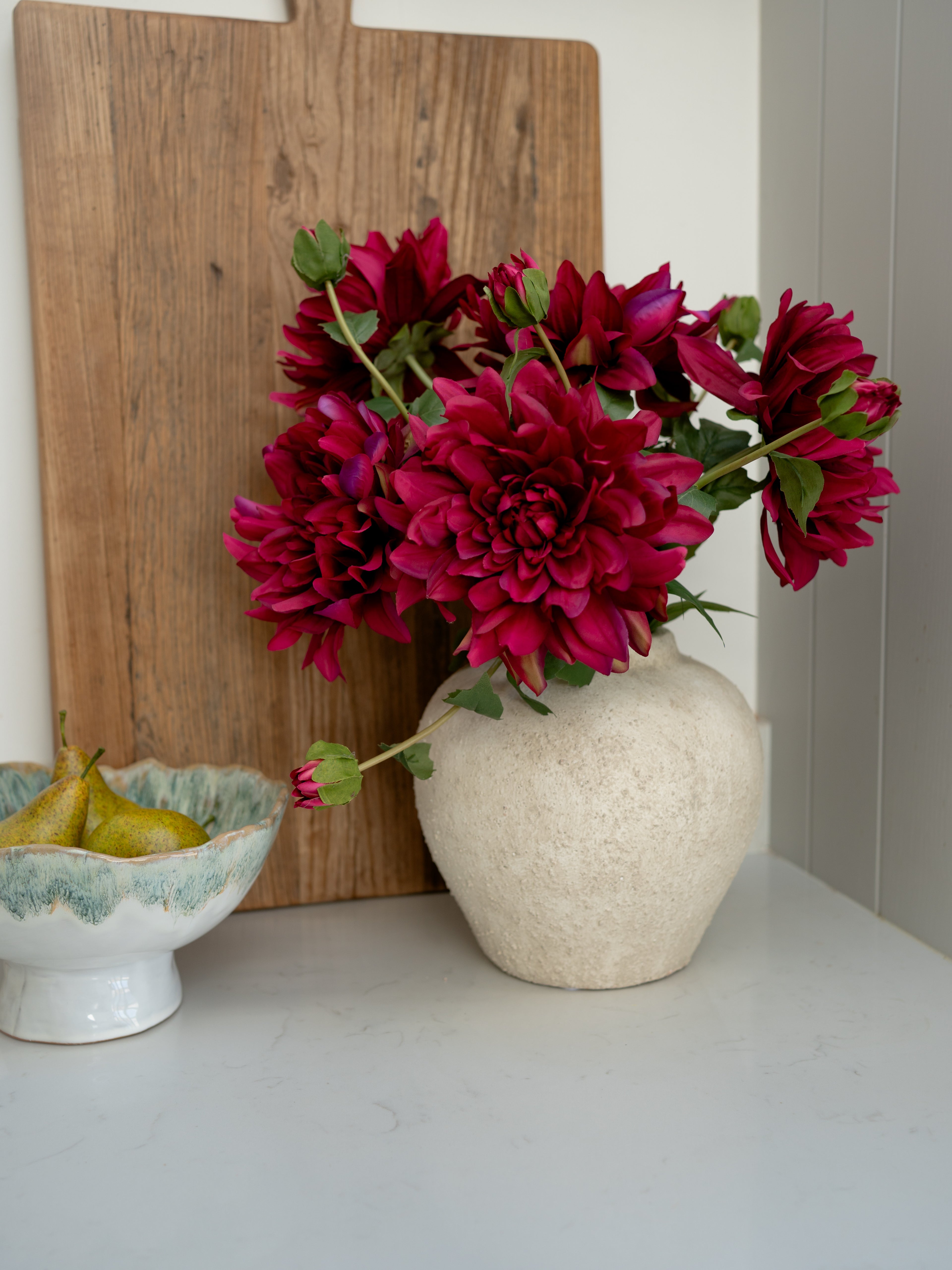 Vase with red flowers on a white surface next to a wooden cutting board and ceramic bowl.