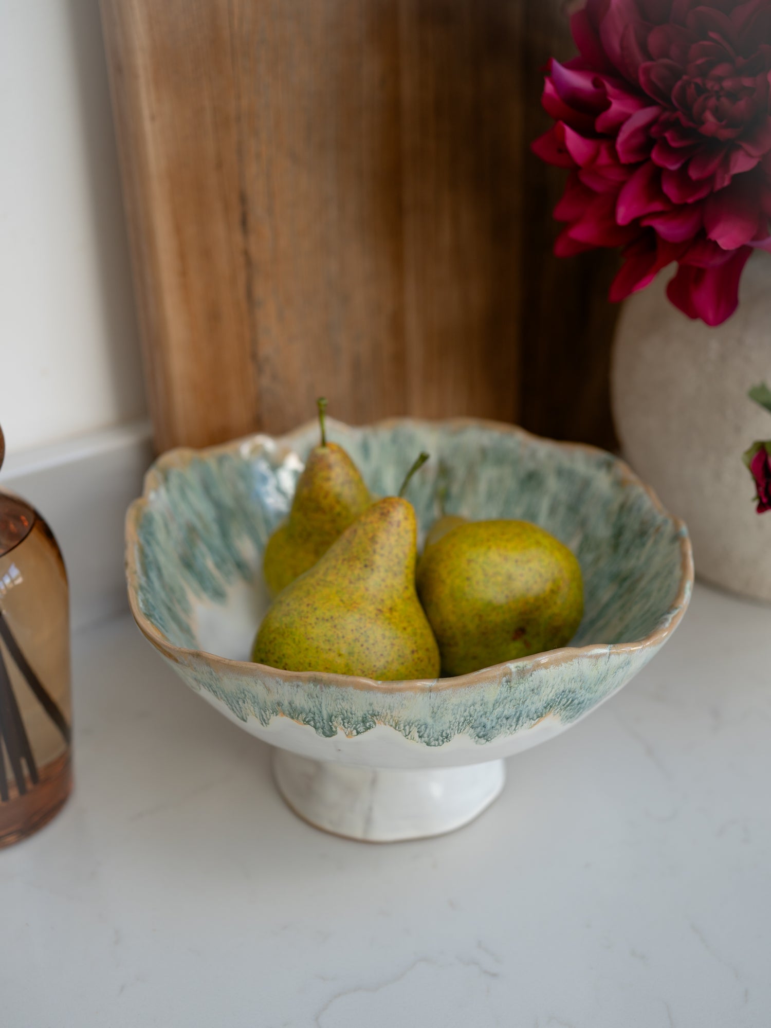 Three pears in a decorative ceramic bowl on a white surface with a wooden cutting board and pink flowers in the background.