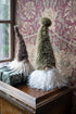 Two fuzzy Christmas gnome decorations displayed on a wooden chest against a floral wallpaper background. One gnome has a tall olive-green hat and white beard, while the other has a brown hat and cream beard.