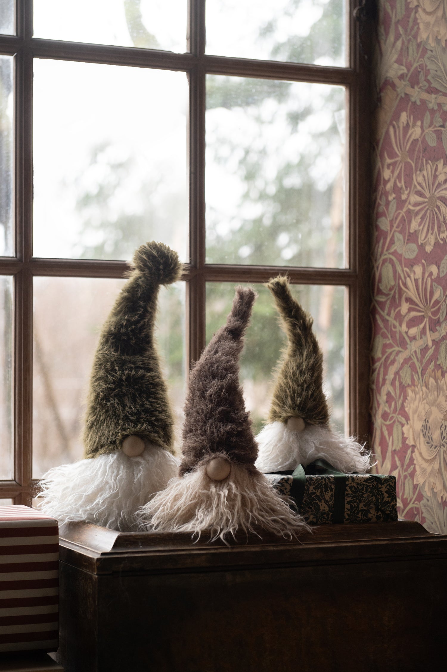 Three fuzzy Christmas gonk decorations with tall pointed hats and white beards sit on a dark wooden chest in front of a window. The gnomes have textured olive green, brown, and tan hats with white and cream beards, and one is posed behind a wrapped gift box.