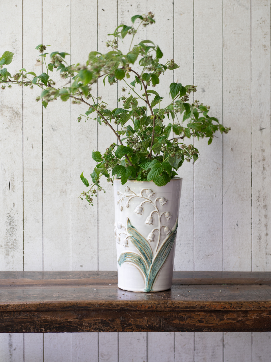 Ceramic vase with embossed lily of the valley design with green plant against a wooden background