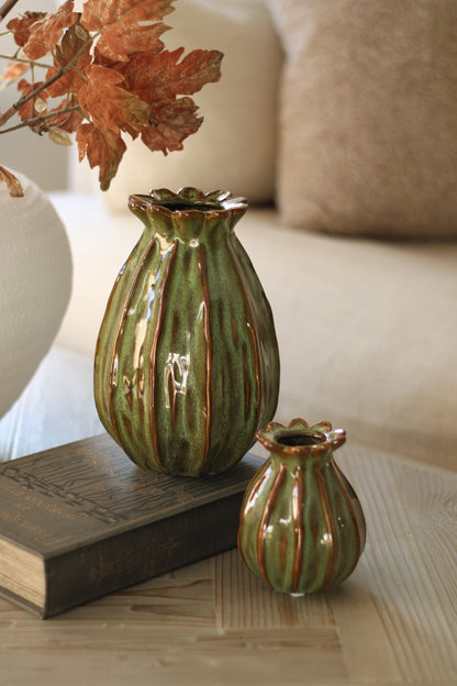 Two green ceramic green  poppy head vases on a surface with a book and decorative leaves.