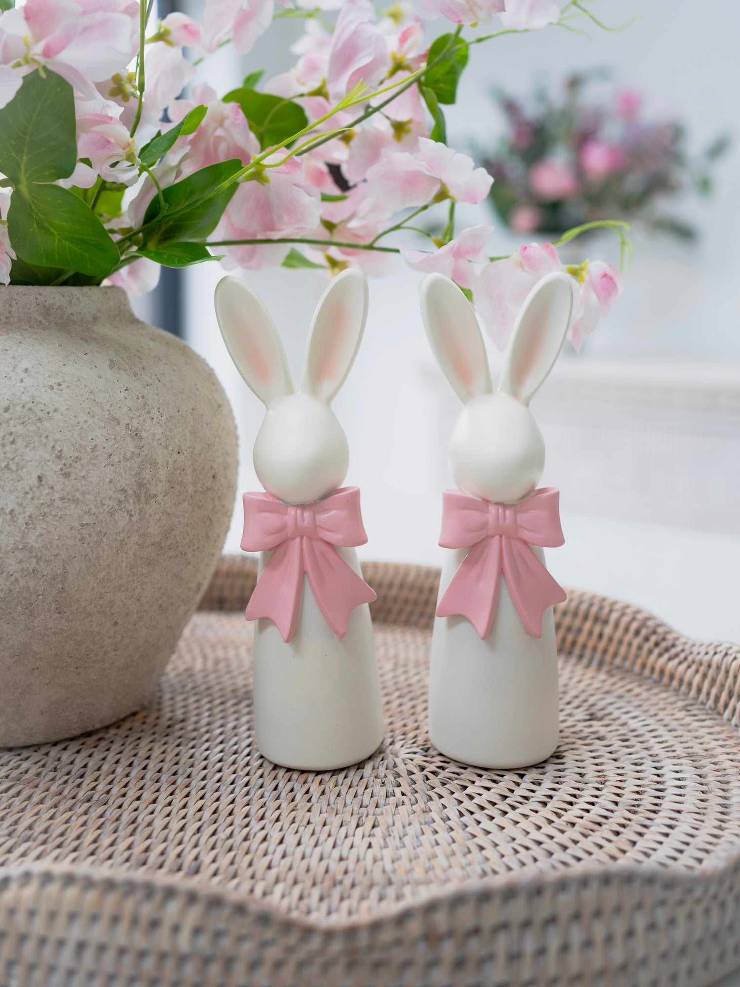 Two white resin bunny figurines with pink bows on a woven surface with a blurred floral background.