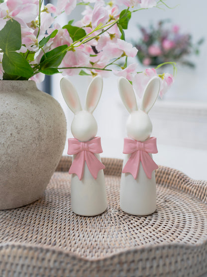 Two white resin bunny figurines with pink bows on a woven surface with a blurred floral background.