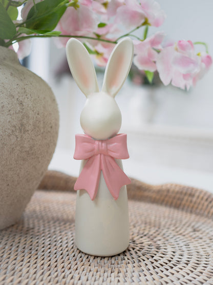 White rabbit figurine with a pink bow on a woven surface with blurred flowers in the background
