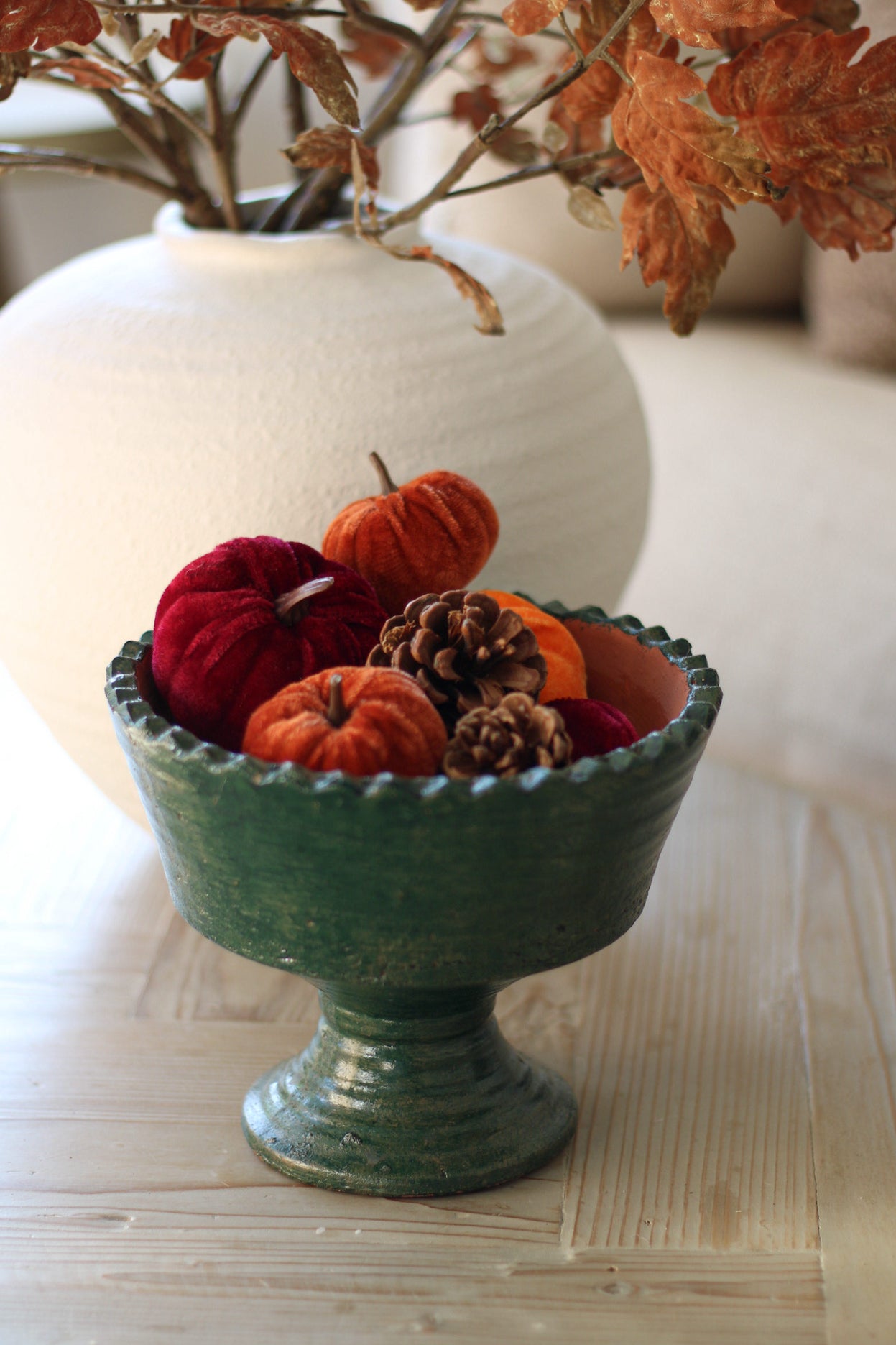 Decorative bowl with pumpkins, pinecones, and berries on a wooden surface with a vase in the background.