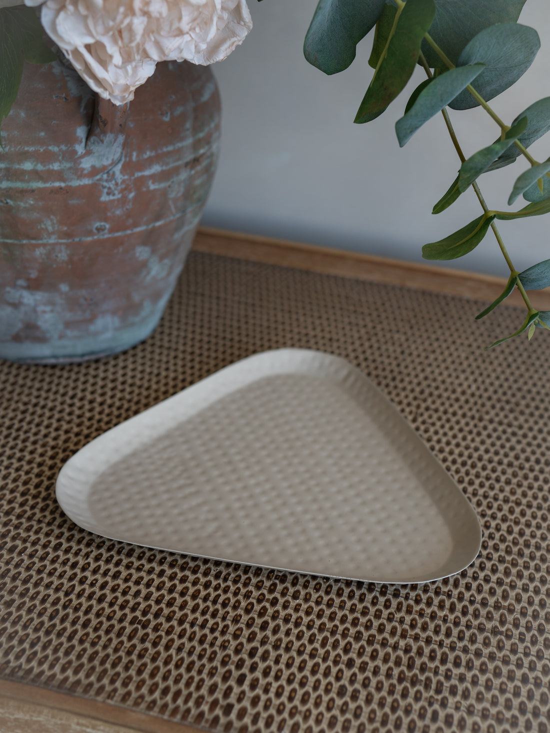 A taupe triangular dimpled metal tray with a textured, dimpled surface, styled on a woven brown placemat with a rustic vase and greenery in the background.