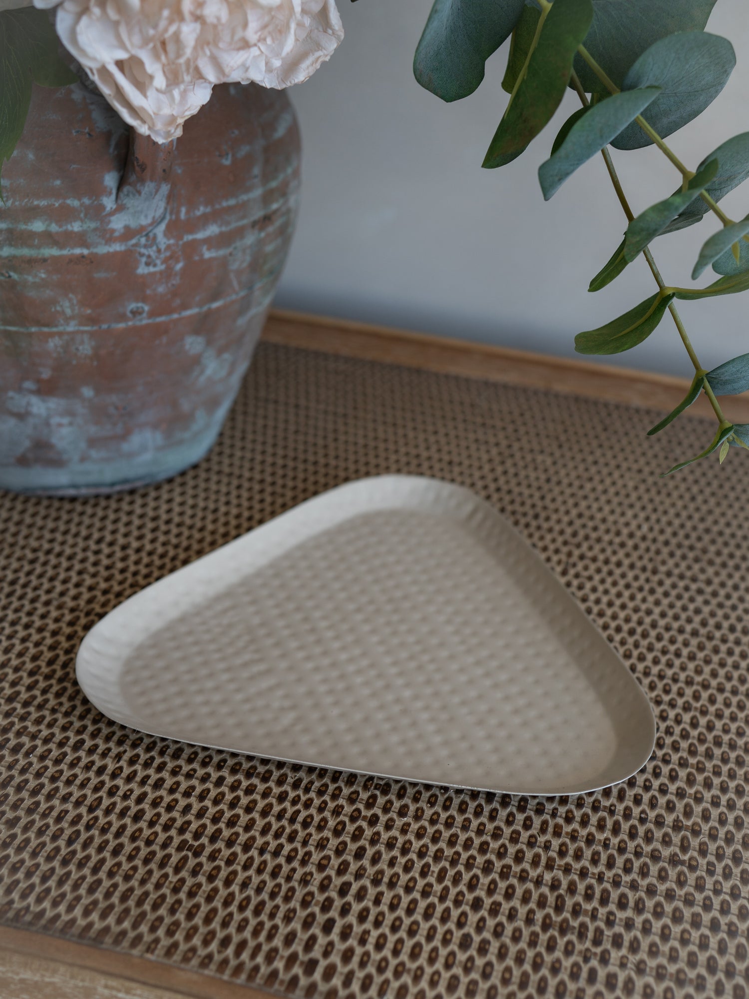 A taupe triangular dimpled metal tray with a textured, dimpled surface, styled on a woven brown placemat with a rustic vase and greenery in the background.