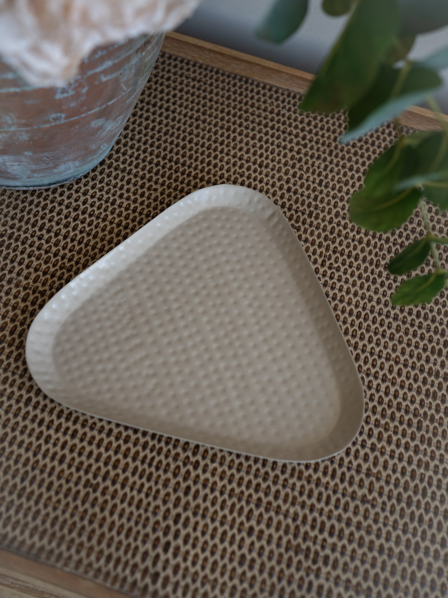 A triangular taupe metal tray with a textured honeycomb pattern, resting on a brown woven placemat with a glass vase and green leaves in the background.
