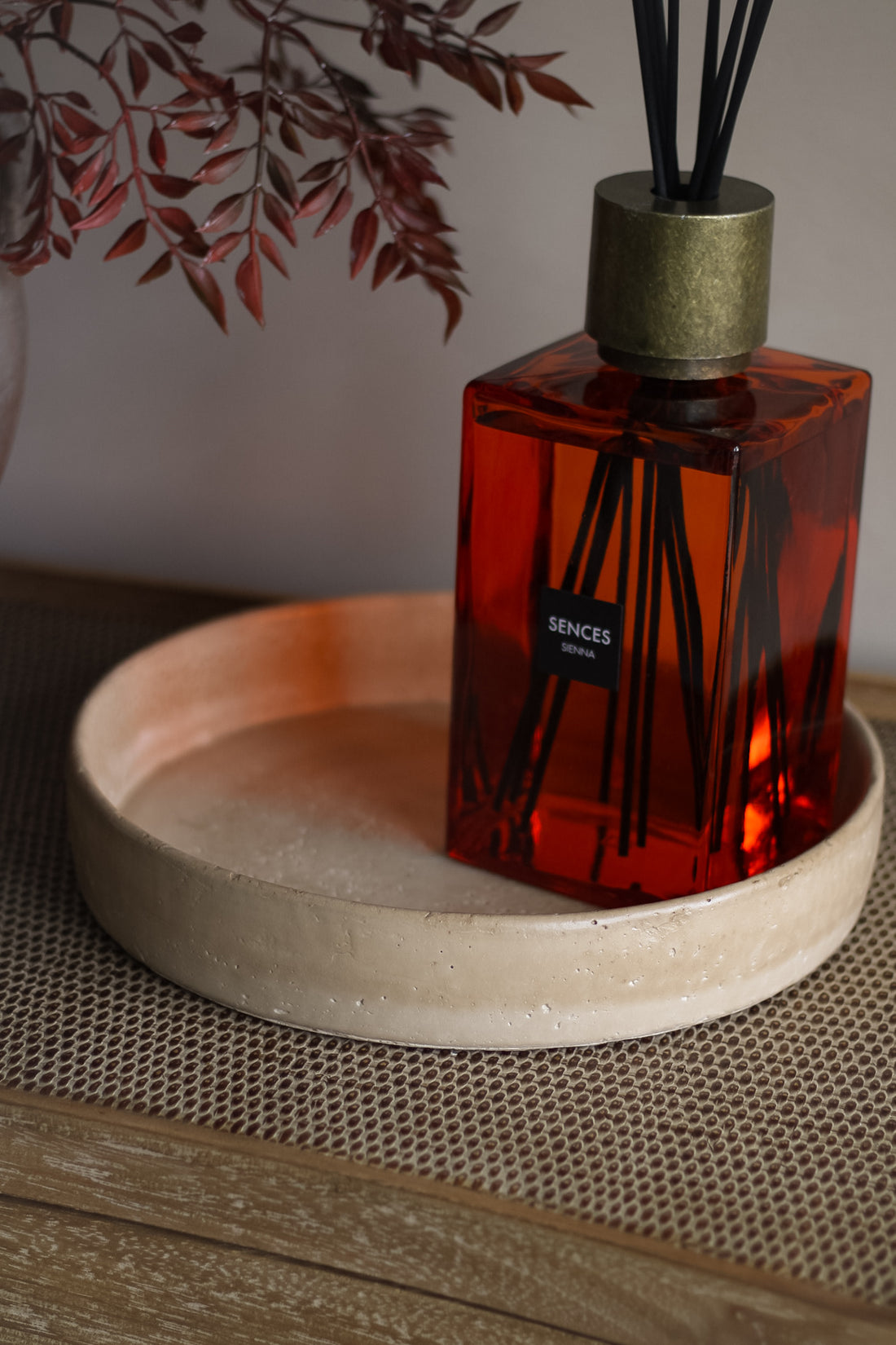 Red diffuser bottle with metallic cap on a beige tray, with blurred leaves in the background