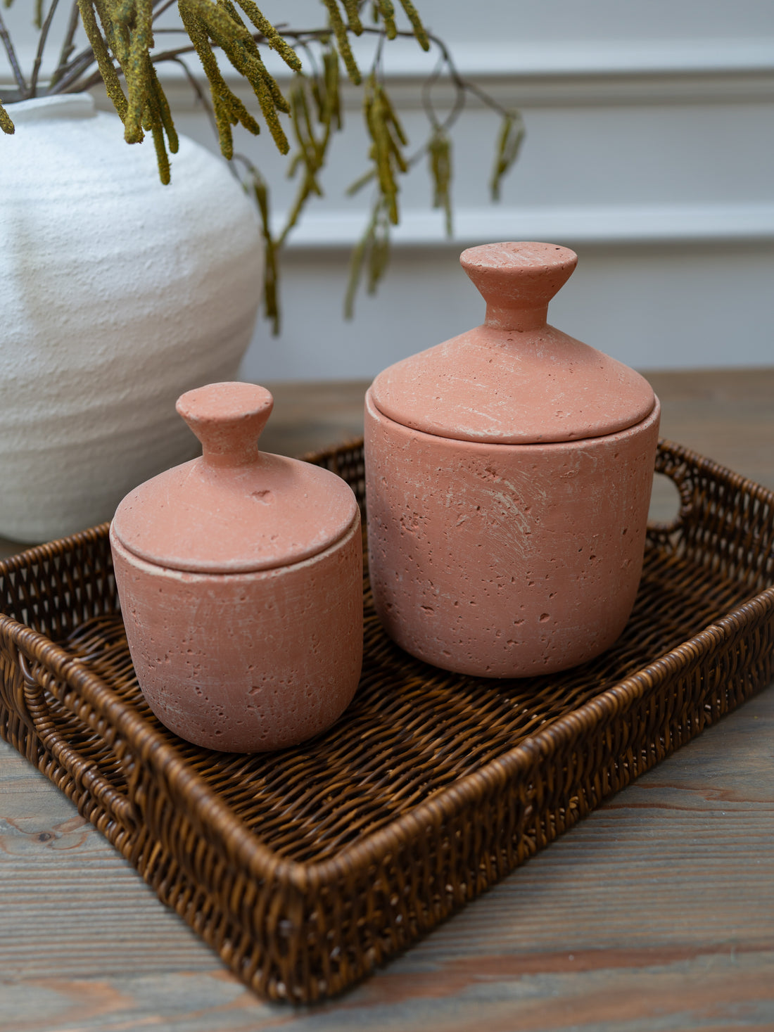 Two terracotta jars with lids on a woven tray with a white vase and plant in the background.