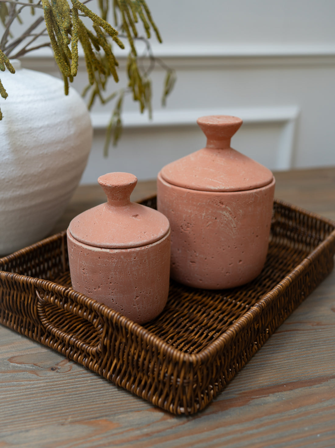 Two terracotta jars with lids on a woven tray with a white vase and greenery in the background.
