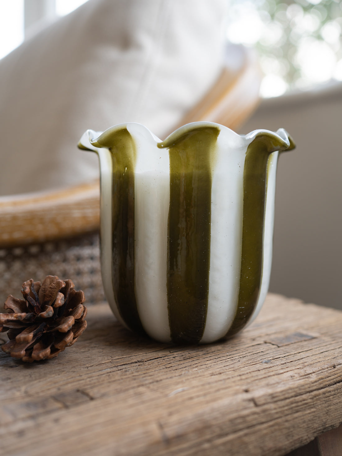 A handmade glass scented candle with vertical olive green and white stripes, styled on a rustic wooden table next to a pinecone.