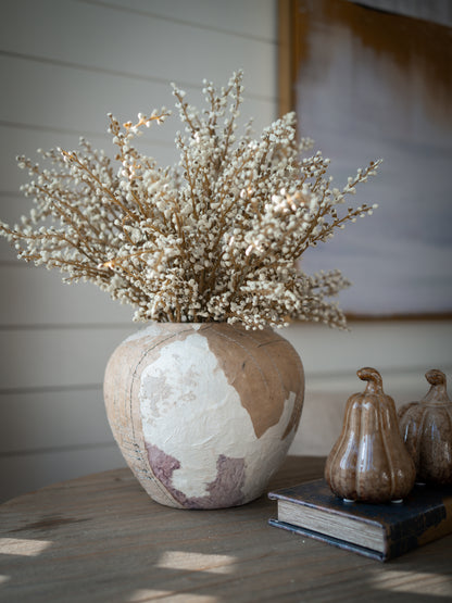 A rustic ceramic vase with a distressed beige, white, and muted mauve glaze, filled with faux soft cream heather spray, displayed on a wooden table next to decorative pumpkins and a book.